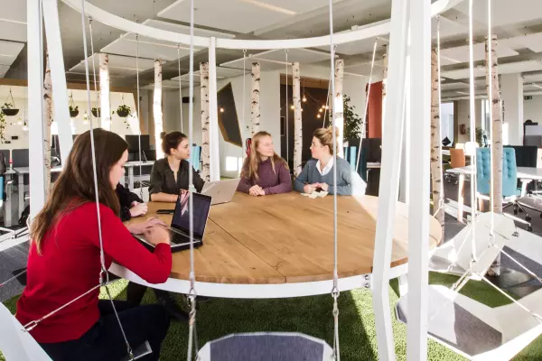 Meeting area with a round wooden table and swing seats in an open-plan office with natural daylight.