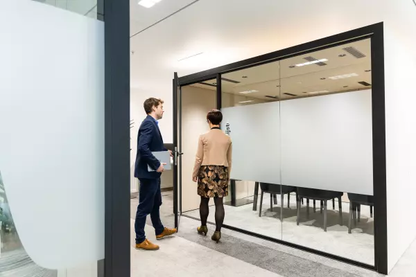 Two colleagues enter a meeting room with glass walls in a modern office.